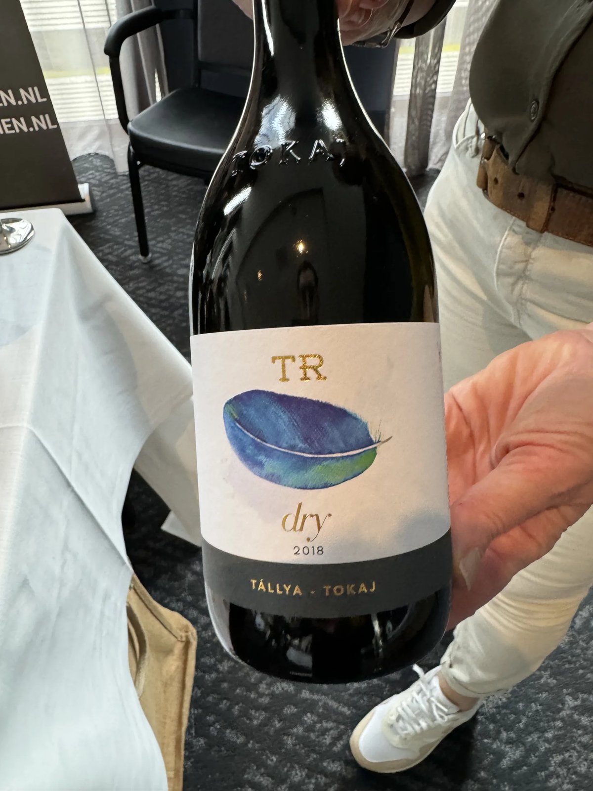 Hand holding a dark Tokaj bottle with a TR dry 2018 label showing a painted blue feather