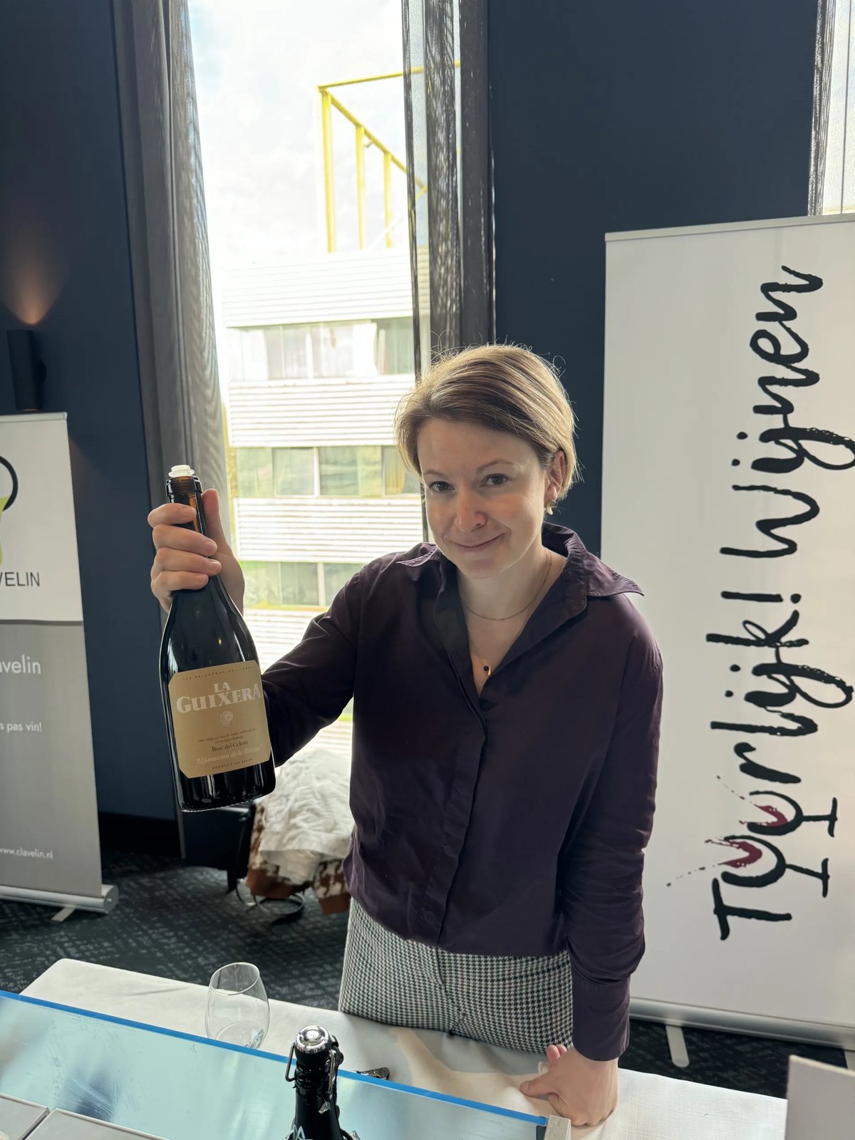 Woman in a purple blouse at the TuurlijkWijnen stand holding a La Guixera bottle by the neck
