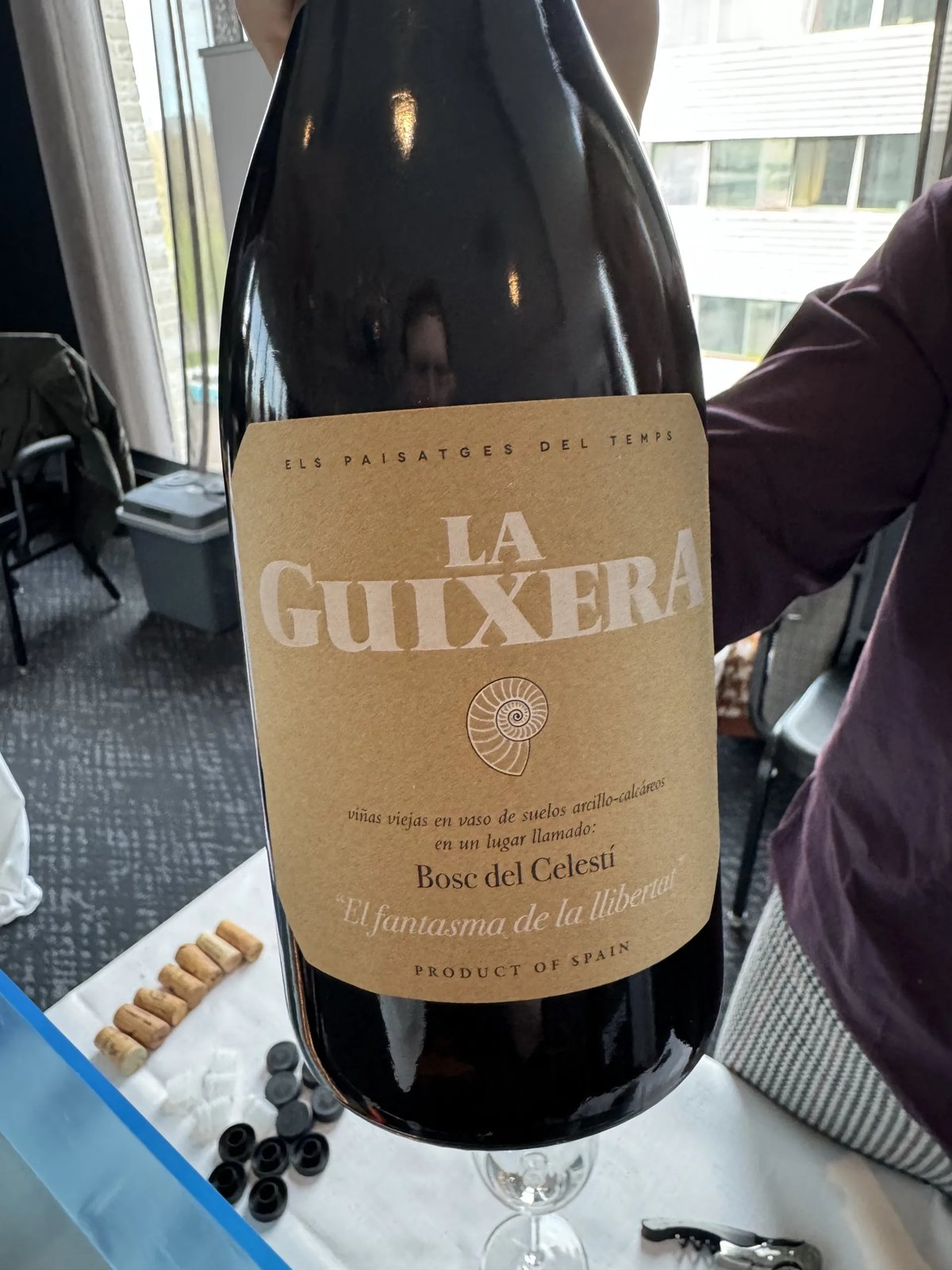 Large dark La Guixera bottle held up close, kraft label reading Els Paisatges del Temps with an ammonite drawing
