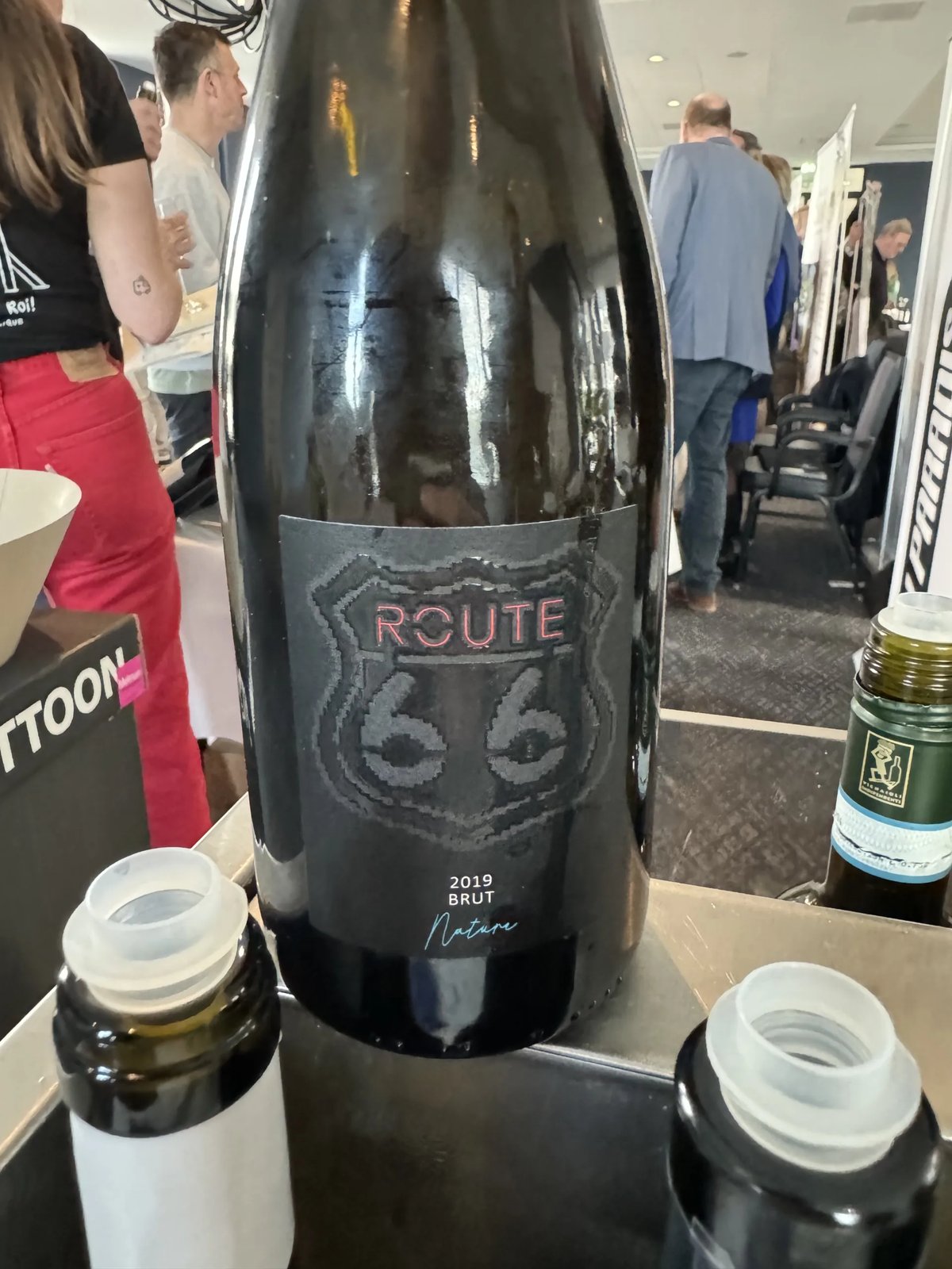 Dark sparkling bottle with a Route 66 brut nature 2019 label, surrounded by other open bottles at the booth