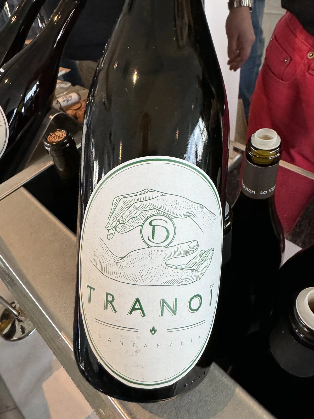 Dark bottle with an oval Tranoï Santamaria label showing a green line drawing of cupped hands