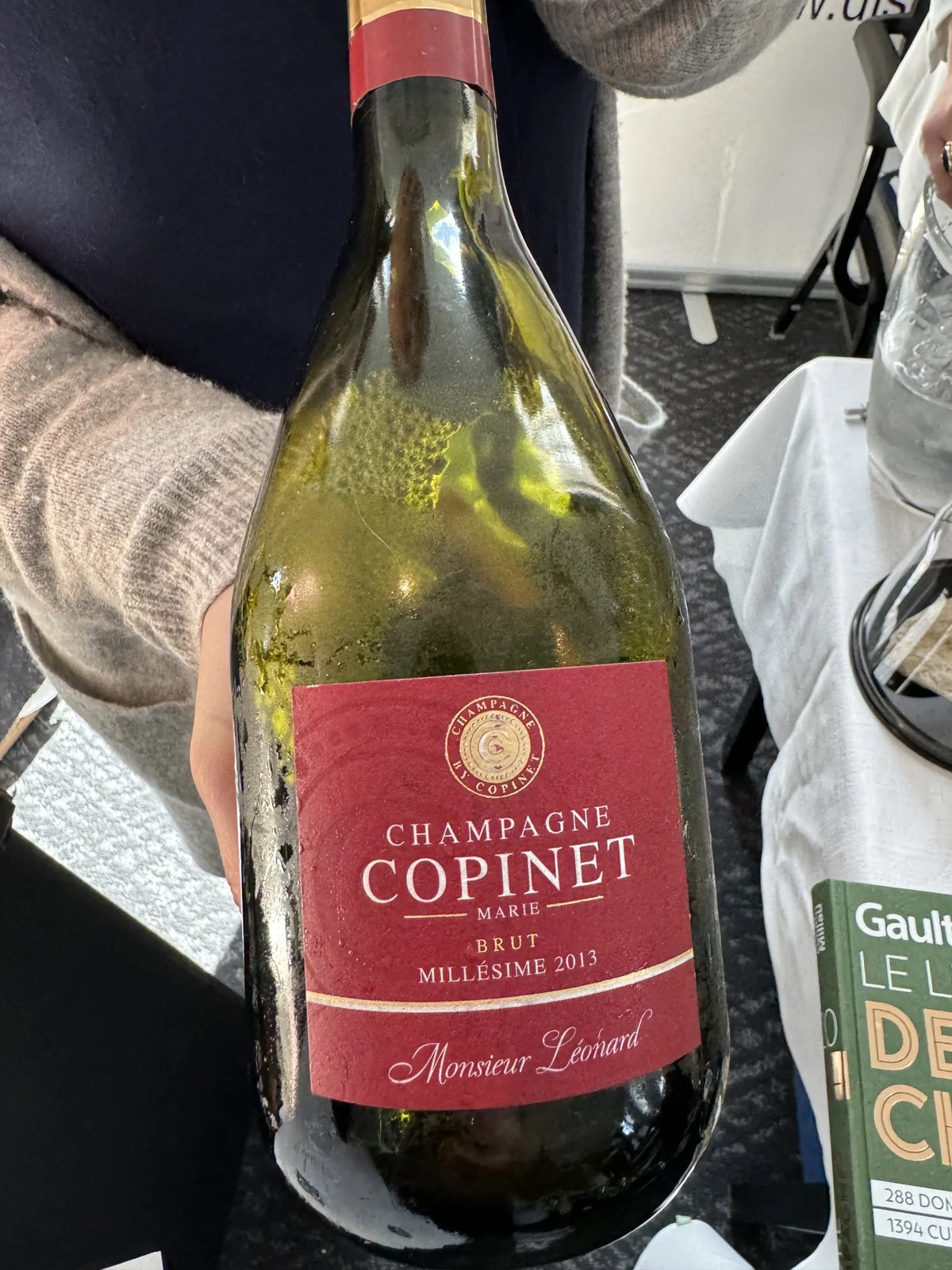 Champagne Marie Copinet Monsieur Léonard Millésime 2013 bottle with a red label, held against a sweater