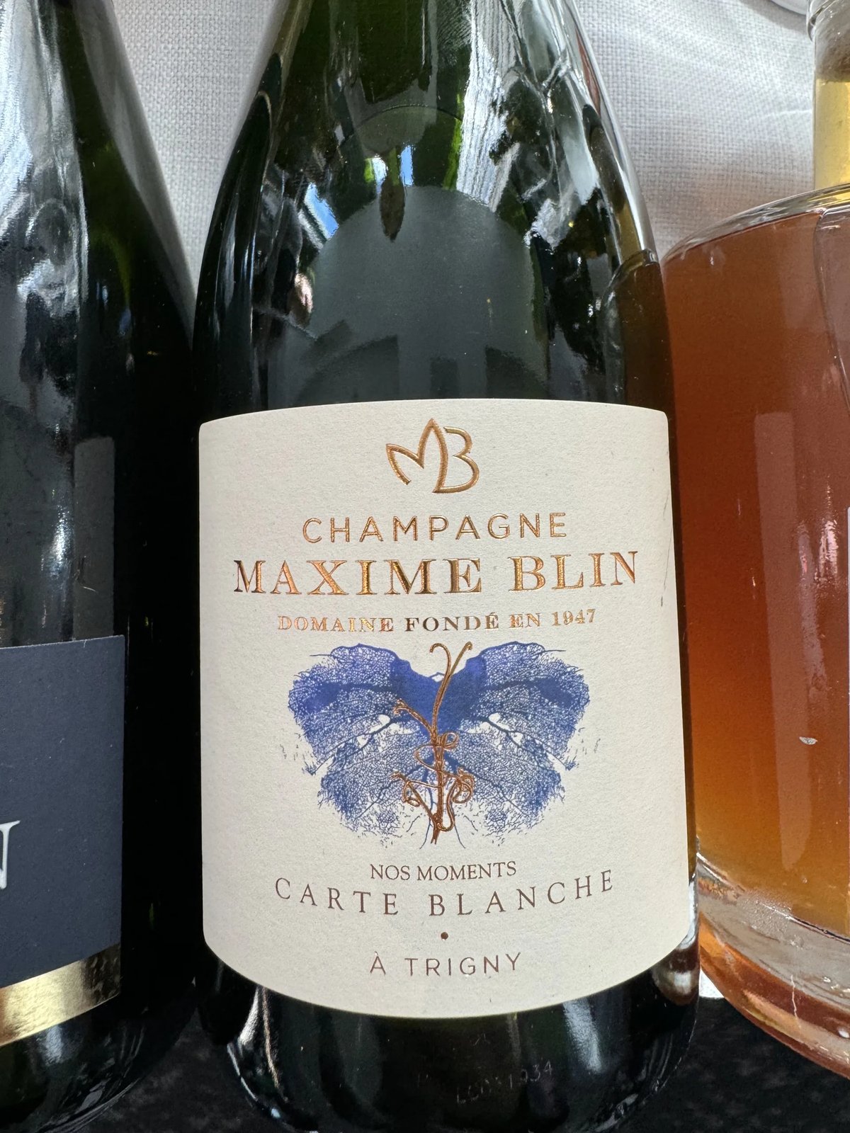 Champagne Maxime Blin Carte Blanche bottle with a cream label and a blue butterfly-shaped vine print