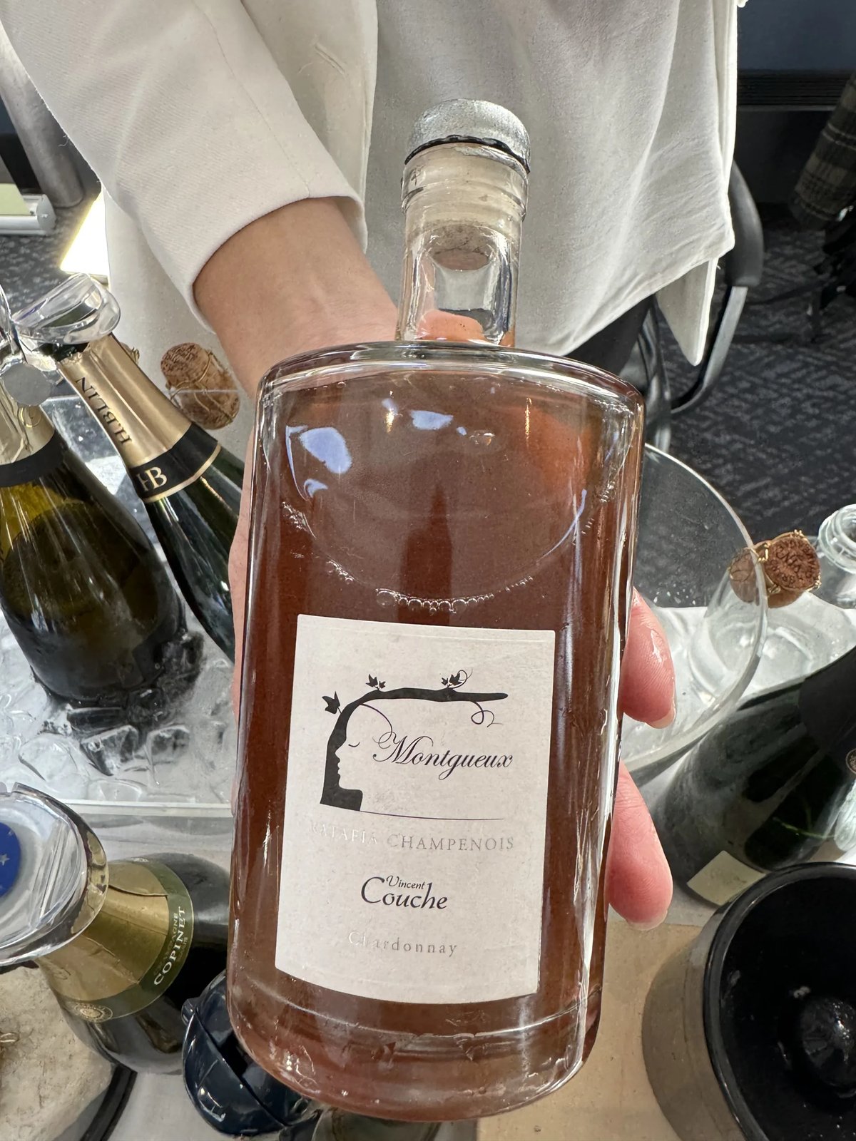 Squat jar-shaped Vincent Couche Montgueux Ratafia bottle of pink liquid, held above ice and champagne bottles