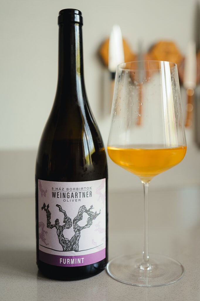 Weingartner Furmint bottle with a purple vine-drawing label beside a glass of deep amber-orange wine