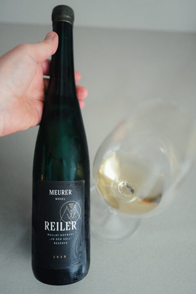 Hand holding a dark green Meurer Reiler Reserve 2020 bottle beside a glass of pale white wine