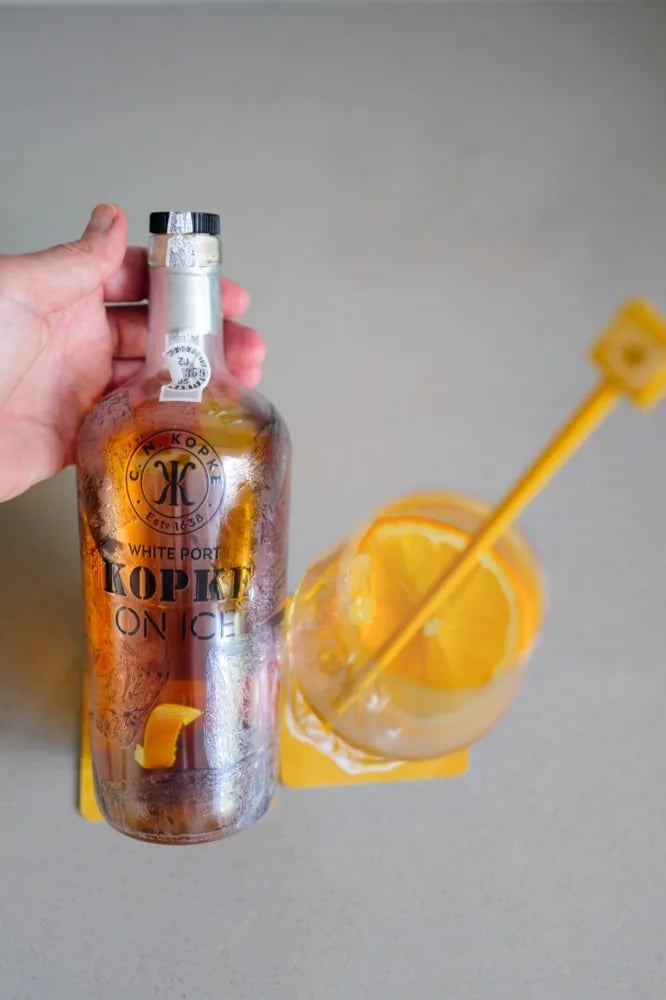Hand holding a glass of Kopke On Ice with ice and a slice of orange next to the bottle