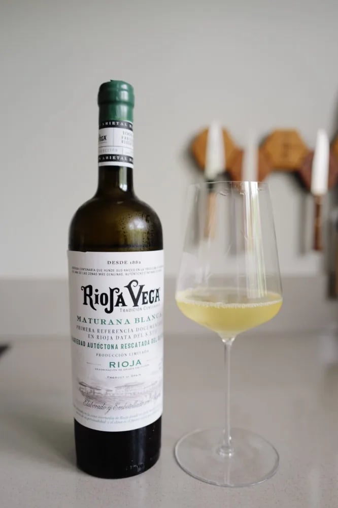 Bottle of Rioja Vega Maturana Blanca 2025 with front label next to a glass of pale golden wine on a kitchen counter