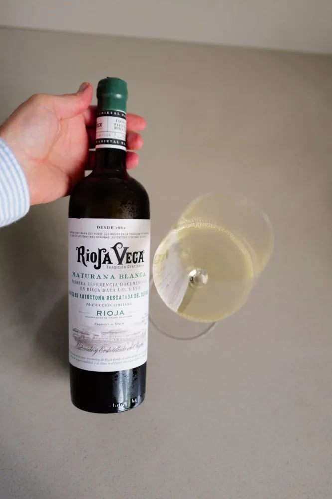 Hand holding the bottle of Rioja Vega Maturana Blanca 2025 next to a tilted glass of white wine