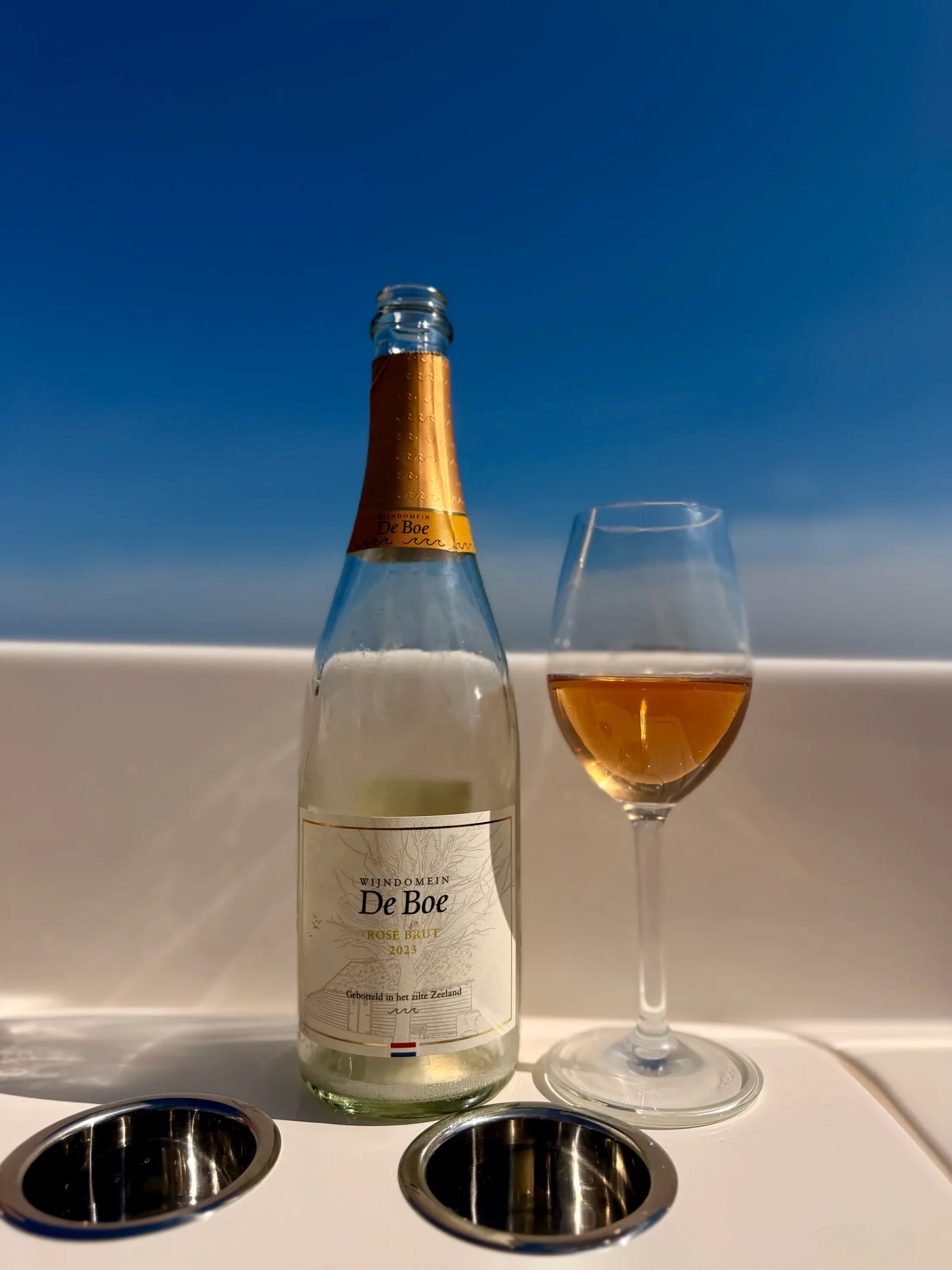 Bottle of Wijndomein De Boe Rosé Brut 2023 beside a glass of copper-coloured sparkling wine on a boat, the water of the Oosterschelde behind it