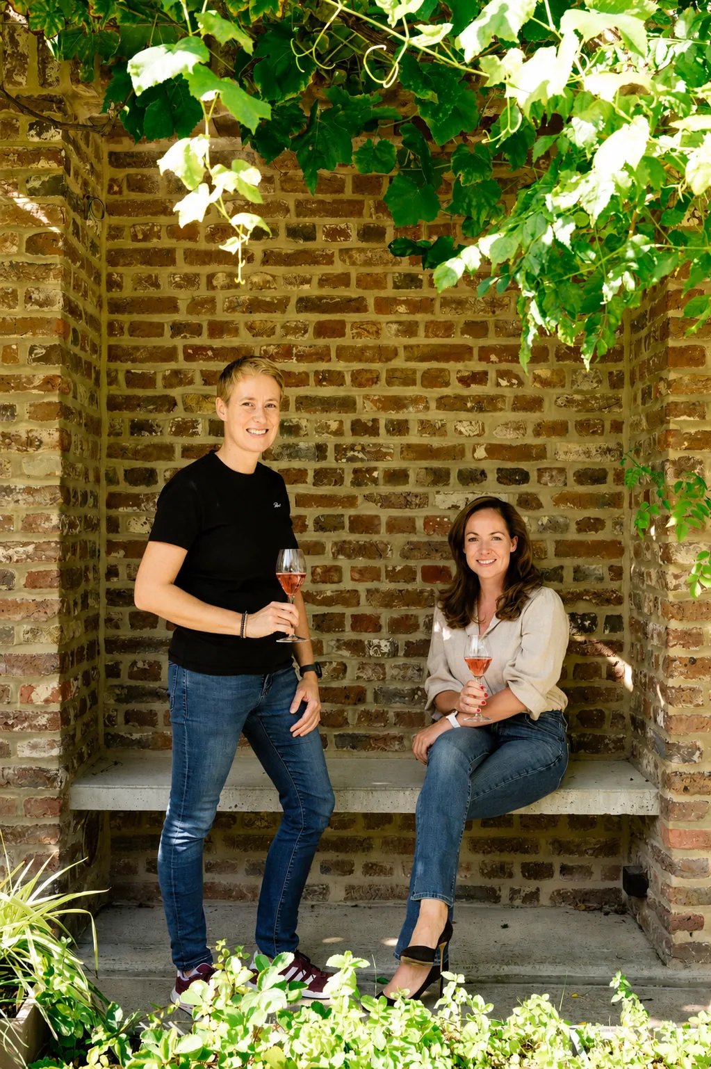 Bottle of Domein Holset Rosé Brut 2023 beside a glass of salmon-pink sparkling wine on a wooden table, greenery and brick behind it
