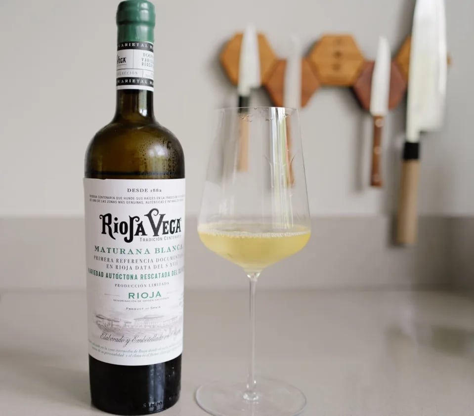 Bottle of Rioja Vega Maturana Blanca 2025 with a glass of white wine on a kitchen counter, Japanese knives on the wall
