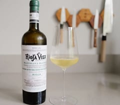 Bottle of Rioja Vega Maturana Blanca 2025 with a glass of white wine on a kitchen counter, Japanese knives on the wall