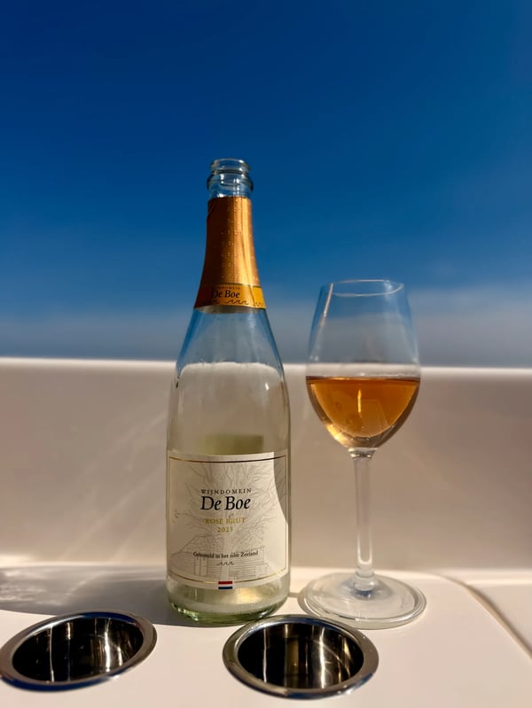 Bottle of Wijndomein De Boe Rosé Brut 2023 beside a glass of copper-coloured sparkling wine on a boat, the water of the Oosterschelde behind it