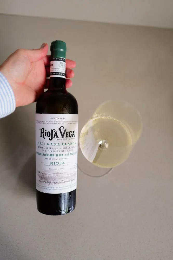 Hand houdt de fles Rioja Vega Maturana Blanca 2025 vast naast een gekanteld glas witte wijn